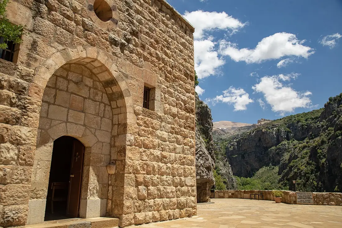 The old stone church of Qannoubine monastery under a bright sky, with the cliffs of the Qadisha Valley behind it. Photo by Hussein Sabboury, Wikimedia Commons