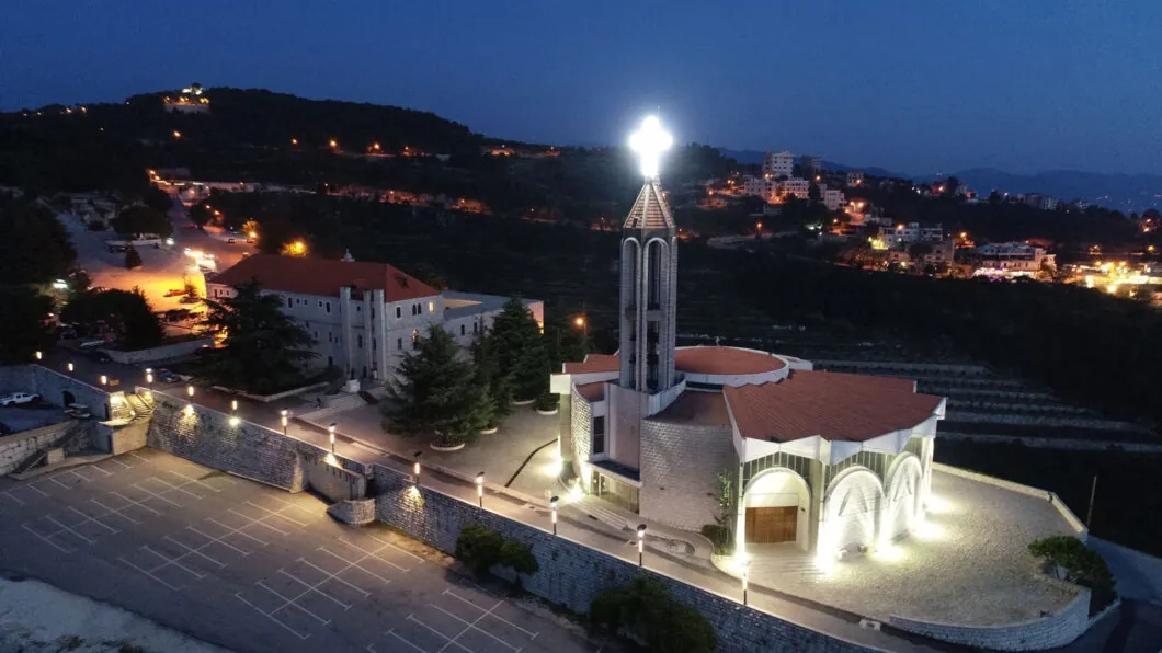 Night view from above of the Annaya church with its lit cross and the monastery buildings among the pines and village lights